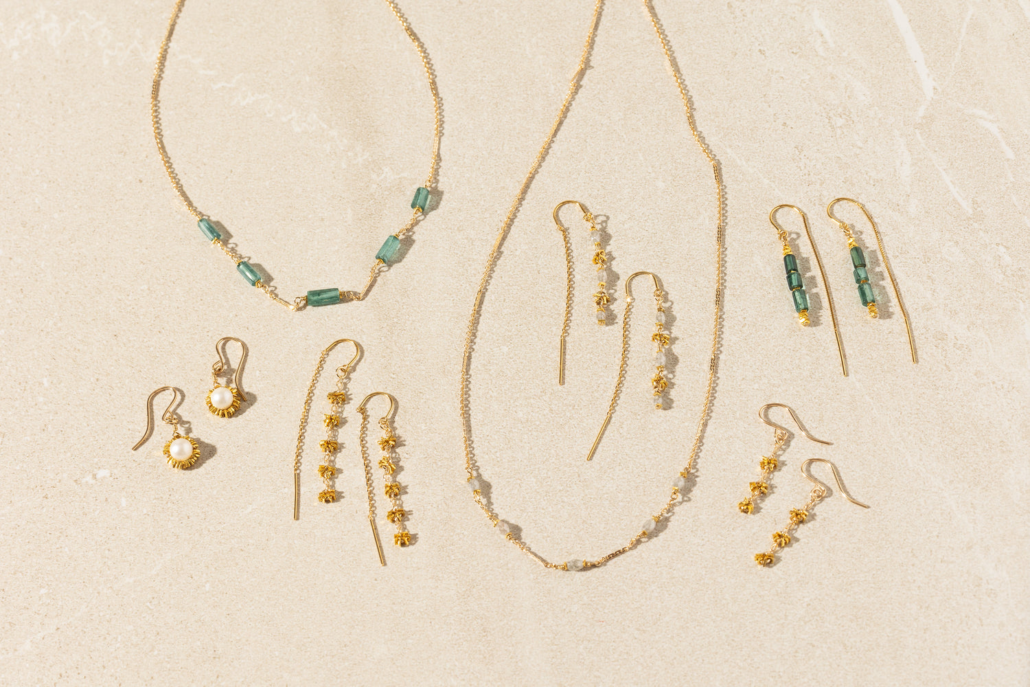 A coordinated jewelry set laid out on a light beige textured surface, including gold-tone necklaces and multiple pairs of drop earrings with green rectangular beads, white pearl-like accents, and small gold beads.