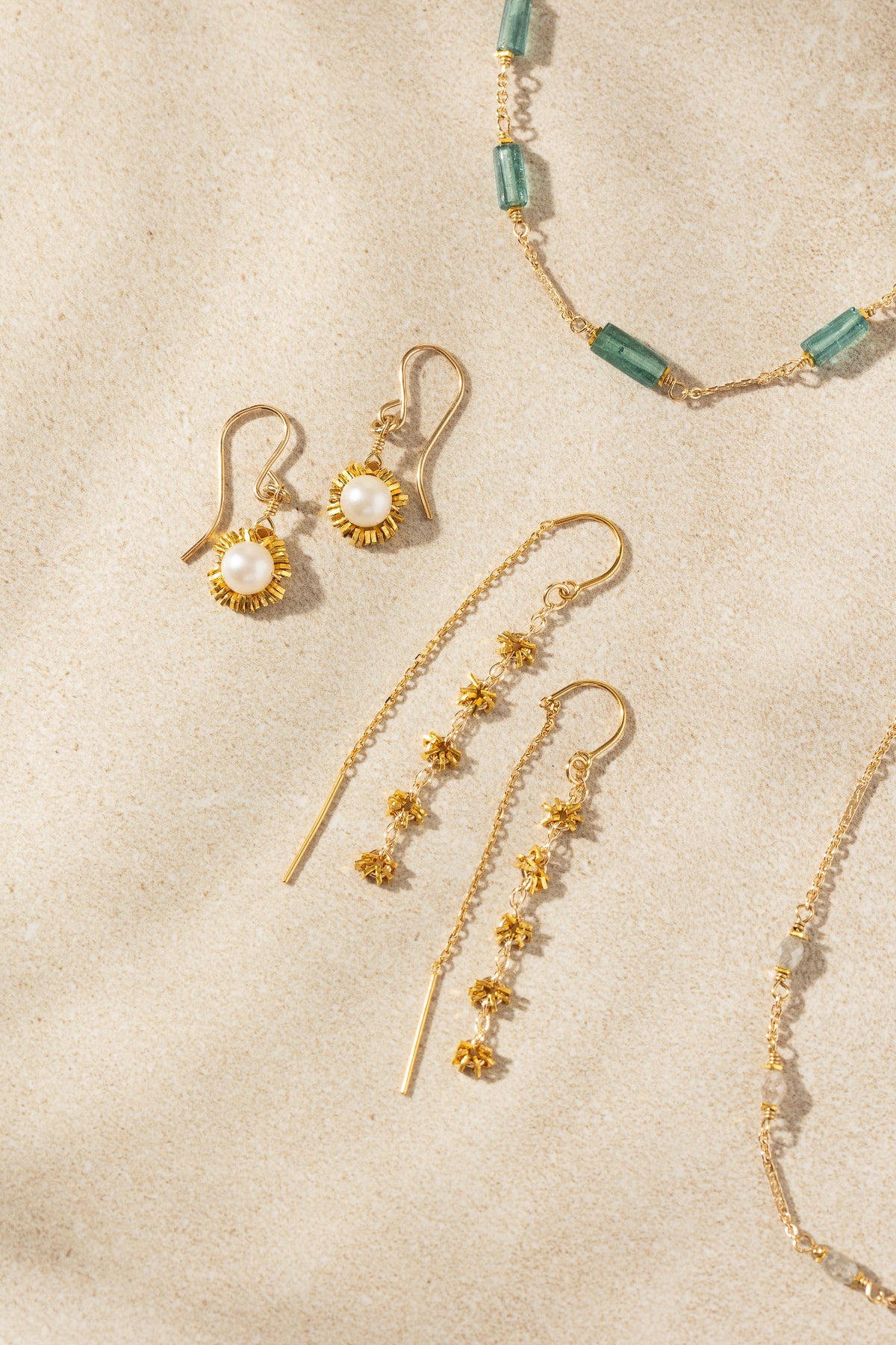 Gold-toned jewelry set including dangle earrings, a necklace, and a bracelet arranged on a textured beige surface. The earrings feature gold floral clusters and pearl-like centers, with a matching beaded necklace in green and gold visible at the top right.