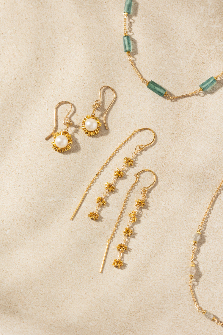 Gold-toned jewelry set including dangle earrings, a necklace, and a bracelet arranged on a textured beige surface. The earrings feature gold floral clusters and pearl-like centers, with a matching beaded necklace in green and gold visible at the top right.