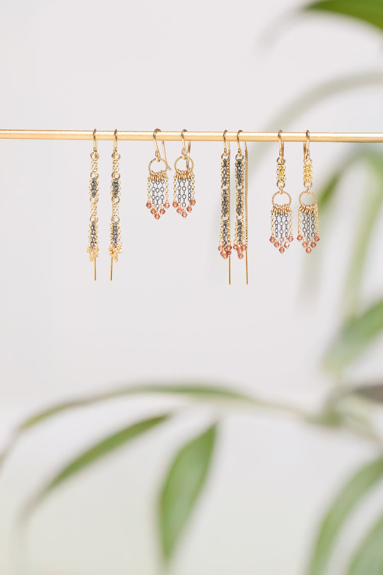 Four pairs of gold and oxidized silver earrings on a gold stand with a blurred green plant background