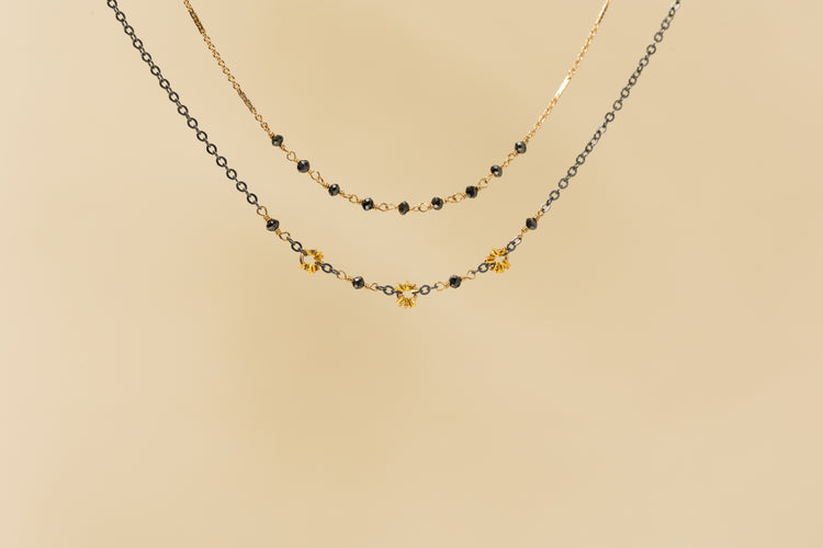 Two delicate layered necklaces displayed against a solid beige background. The top necklace is a fine gold chain, while the bottom necklace features a black chain interspersed with small gold beads and three gold flower-shaped charms.