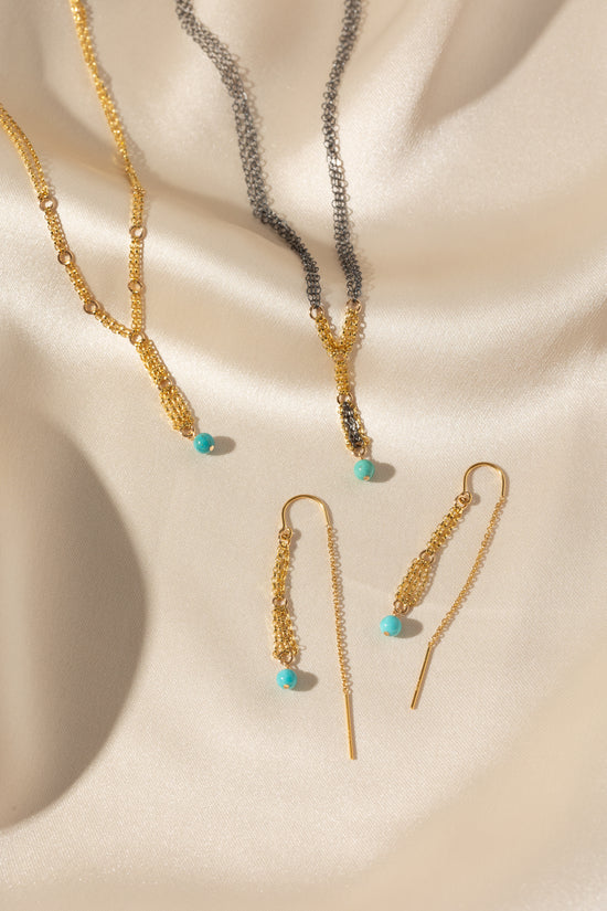 Gold-toned jewelry set including a necklace, matching drop earrings, and a chain bracelet arranged on draped cream satin fabric with turquoise bead accents.