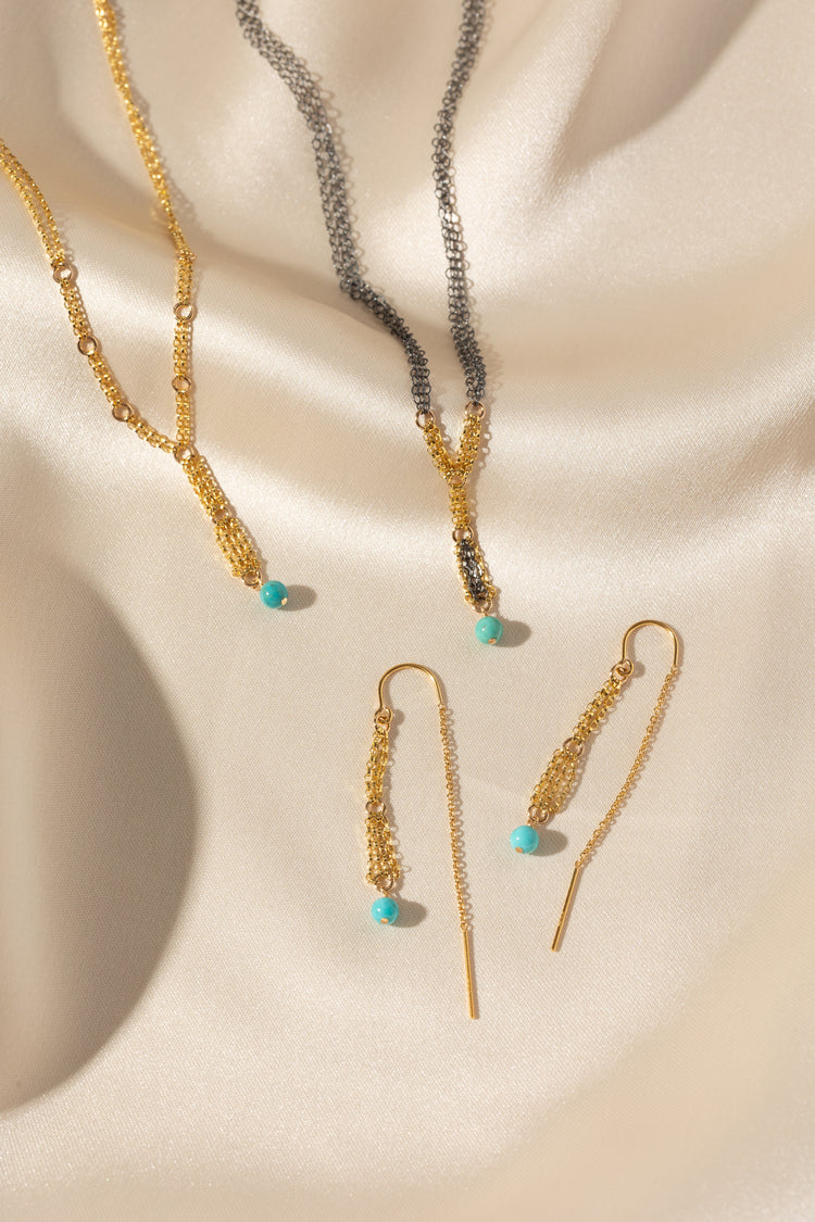 Gold-toned jewelry set including a necklace, matching drop earrings, and a chain bracelet arranged on draped cream satin fabric with turquoise bead accents.