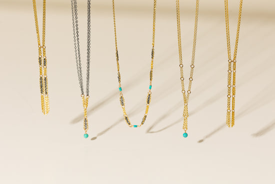 Five delicate gold-toned necklaces with mixed metallic and textured chain details, hanging against a warm cream background. Three of the necklaces feature small turquoise bead accents near the pendants.