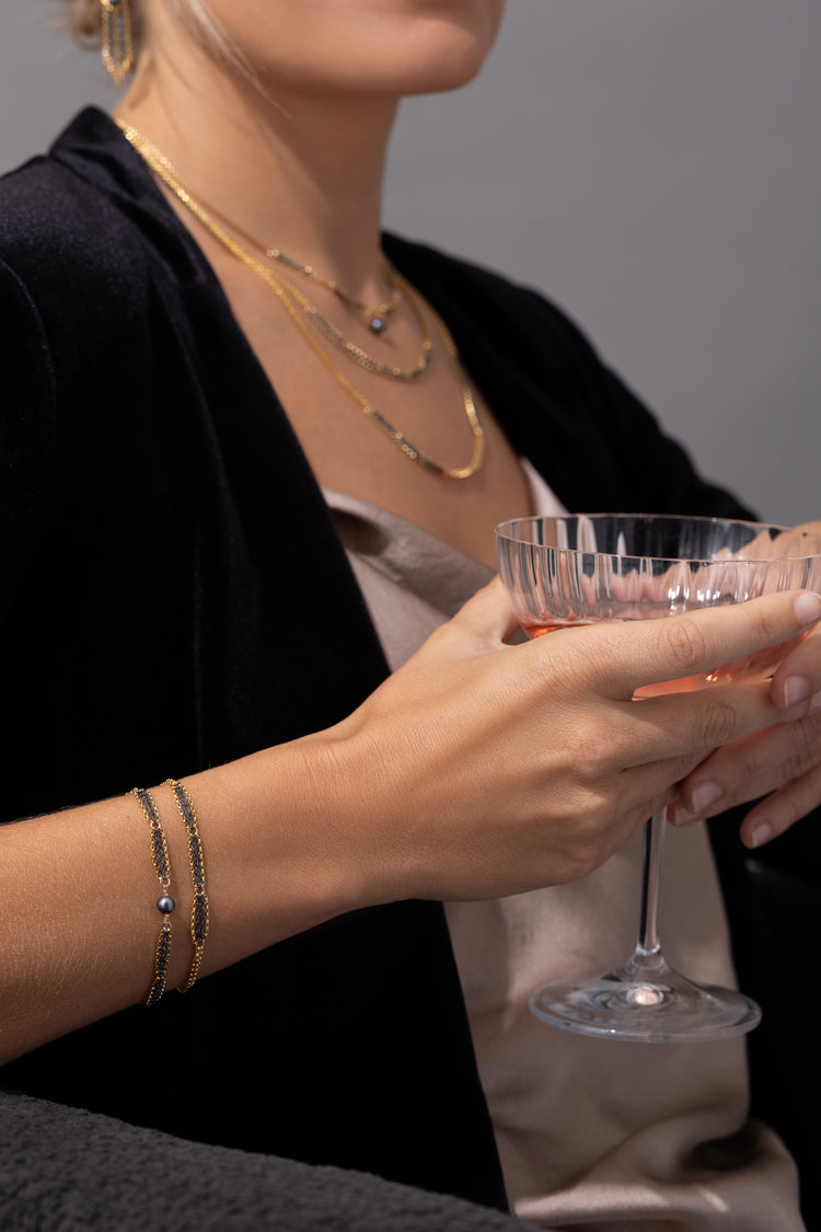 A model holds a drink and is wearing two mixed metal bracelets and layered necklaces in the background 