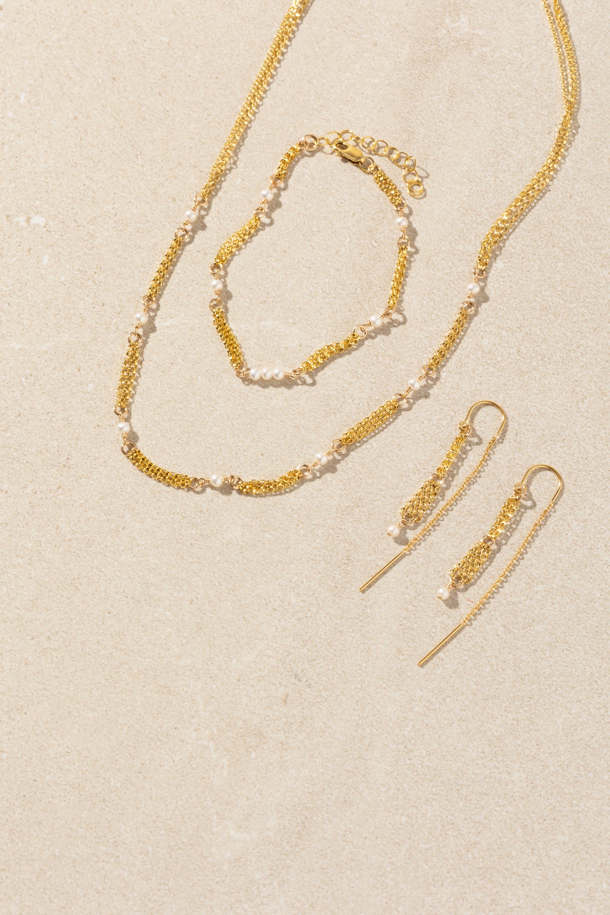 A gold jewelry set with white pearls, including a delicate chain necklace, matching bracelet, and drop earrings, arranged on a textured beige background.