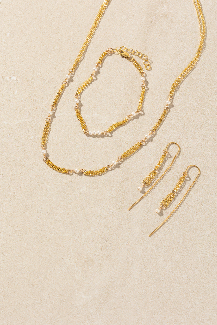 A gold jewelry set with white pearls, including a delicate chain necklace, matching bracelet, and drop earrings, arranged on a textured beige background.