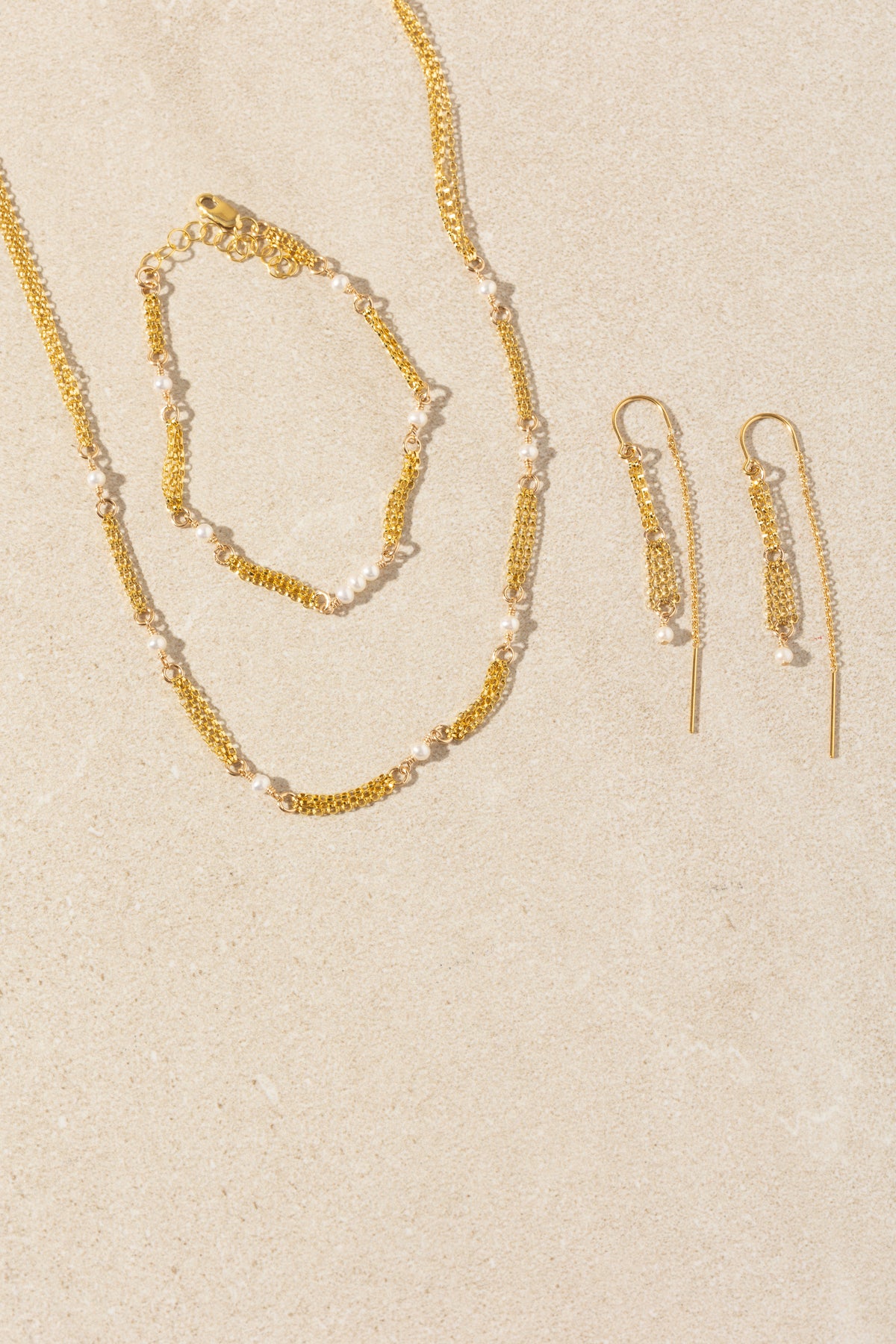A gold jewelry set featuring a delicate chain necklace, matching bracelet, and drop earrings, all embellished with small white pearls, arranged on a textured beige background.