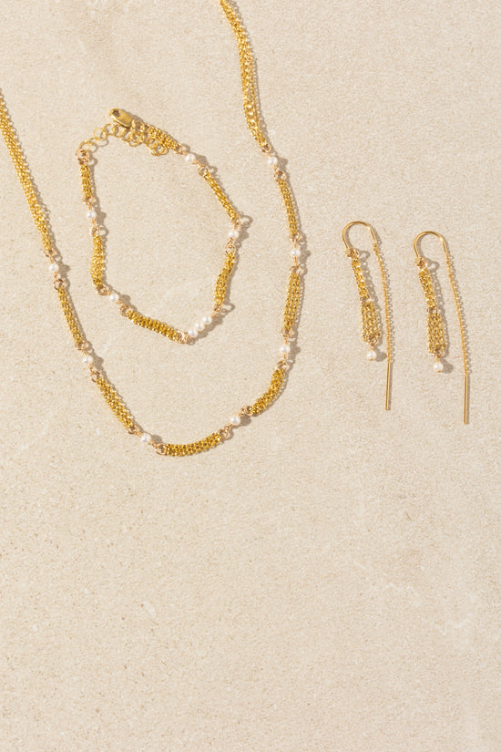 A gold jewelry set featuring a delicate chain necklace, matching bracelet, and drop earrings, all embellished with small white pearls, arranged on a textured beige background.