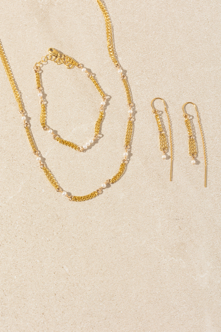 A gold jewelry set featuring a delicate chain necklace, matching bracelet, and drop earrings, all embellished with small white pearls, arranged on a textured beige background.