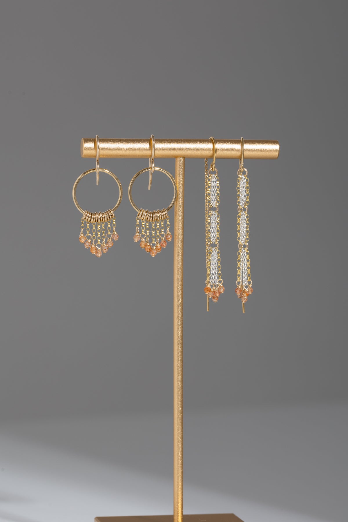 Two pairs of earrings with sunstones hanging on a gold stand with gray background