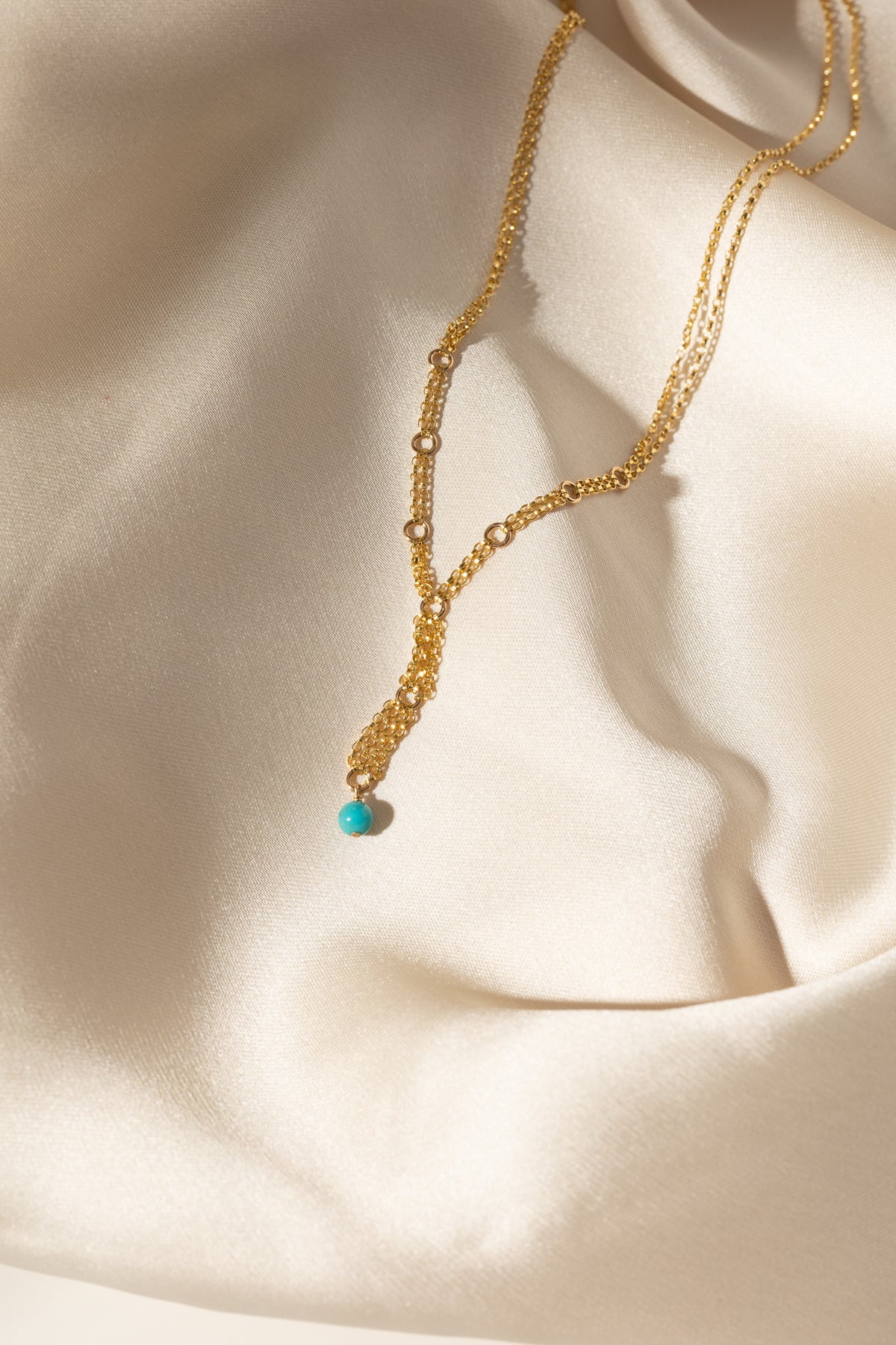 A delicate gold layered necklace with a small turquoise bead pendant, draped elegantly on a cream satin fabric background.