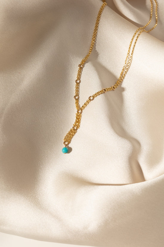 A delicate gold layered necklace with a small turquoise bead pendant, draped elegantly on a cream satin fabric background.