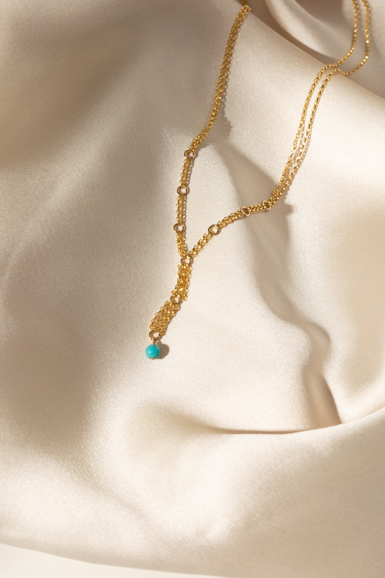 A delicate gold layered necklace with a small turquoise bead pendant, draped elegantly on a cream satin fabric background.