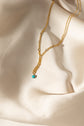A delicate gold layered necklace with a small turquoise bead pendant, draped elegantly on a cream satin fabric background.