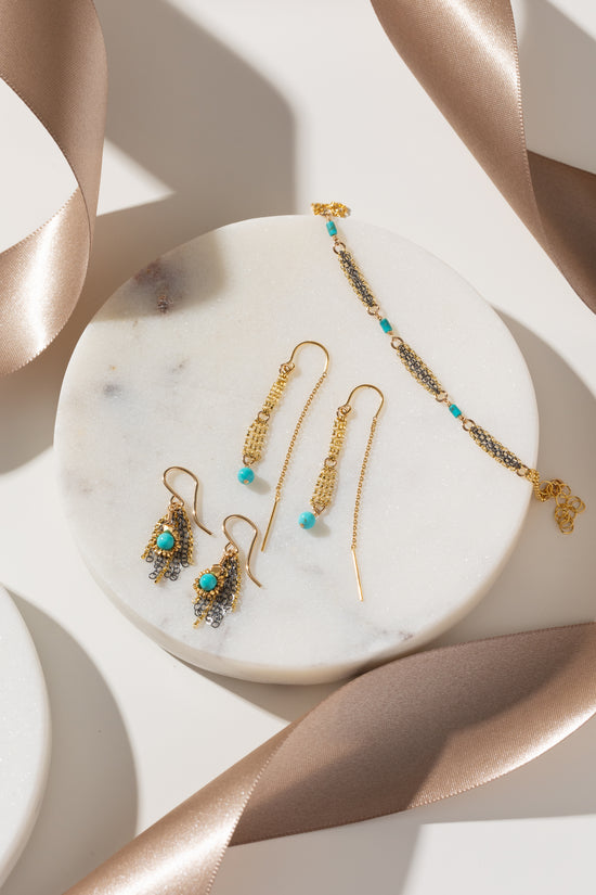 Gold-toned jewelry set including drop earrings and a bracelet with turquoise blue beads, arranged on a round white marble tray with satin ribbon accents.