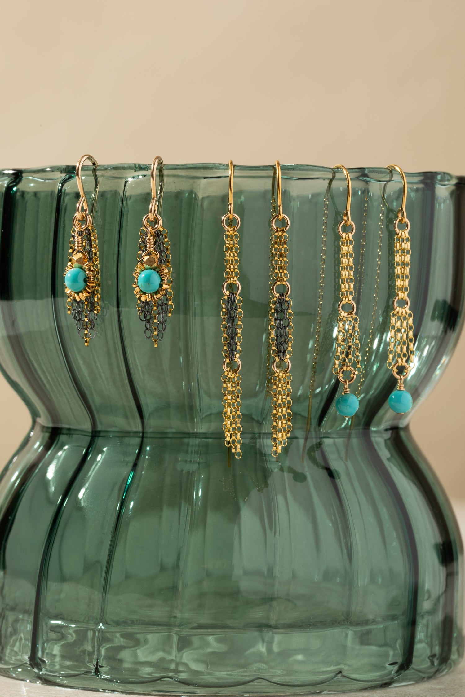 Three pairs of gold-toned dangle earrings with turquoise blue beads and dark gray beaded details, hanging from the rim of a translucent green glass vase against a warm beige background.