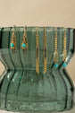 Three pairs of gold-toned dangle earrings with turquoise blue beads and dark gray beaded details, hanging from the rim of a translucent green glass vase against a warm beige background.